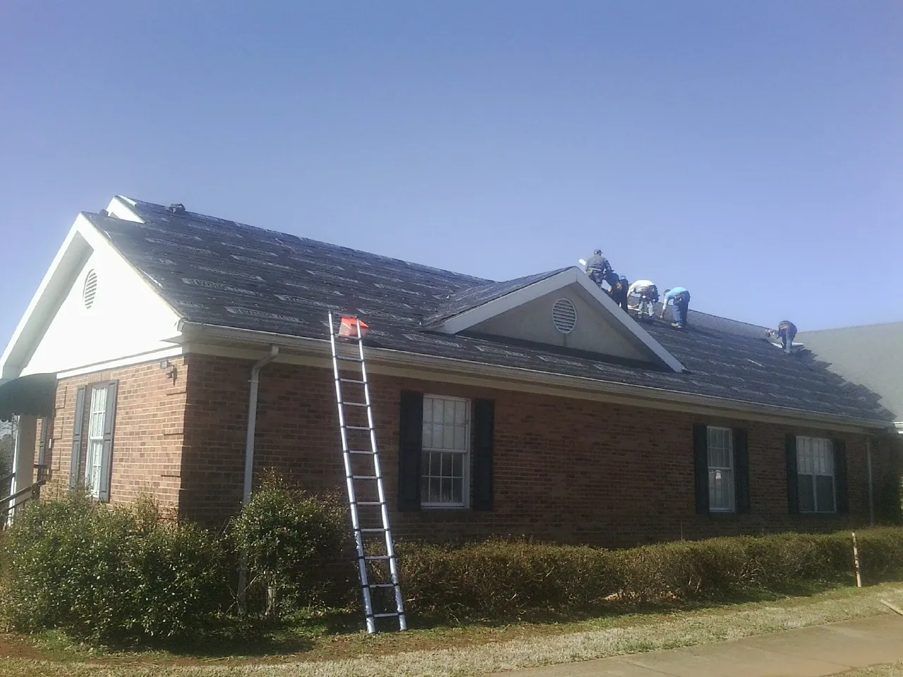 Crew tearing shingles off a brick ranch roof with a ladder set — Emergency Roof Repair work by our crew in Bethpage